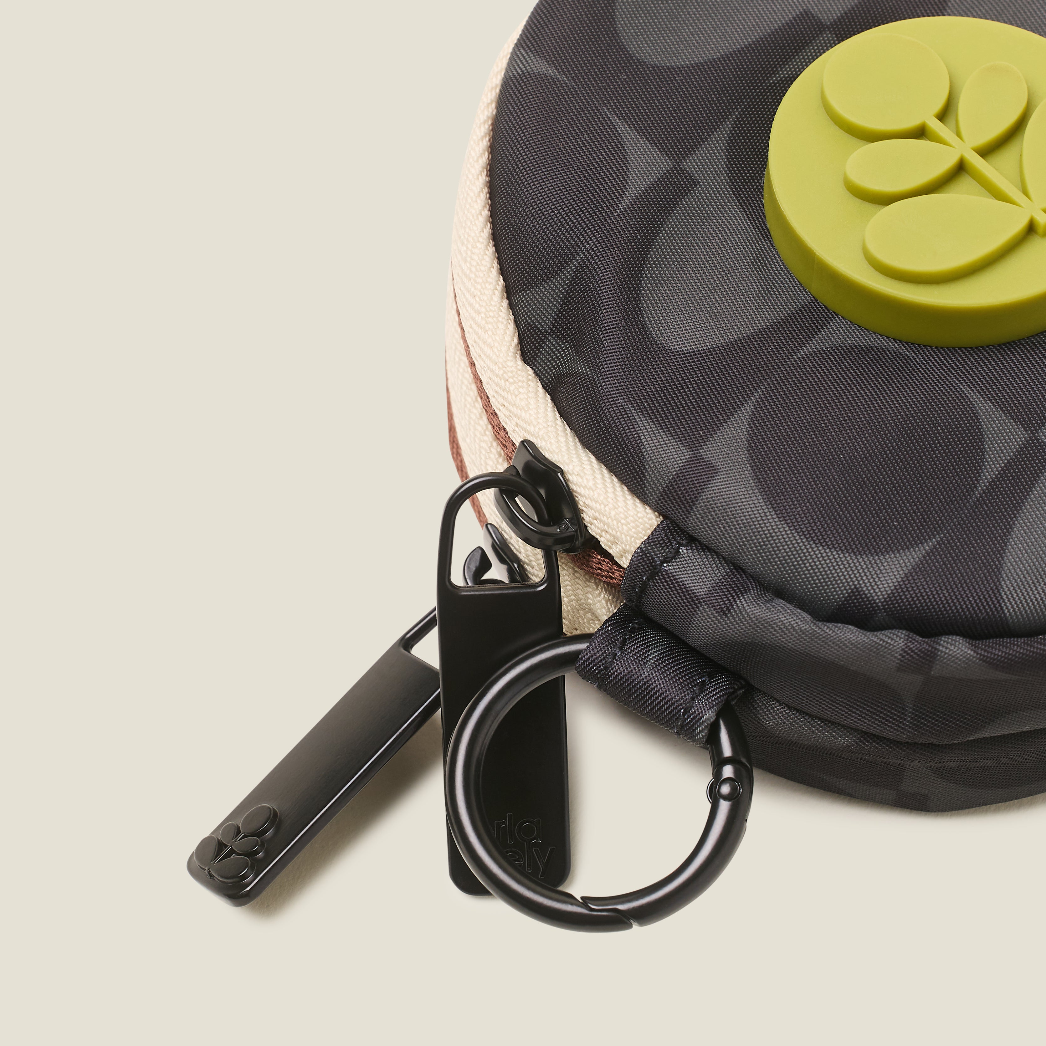 Close-up of a black patterned coin purse with a green rubber logo, branded zip pulls and ring clip on a beige background