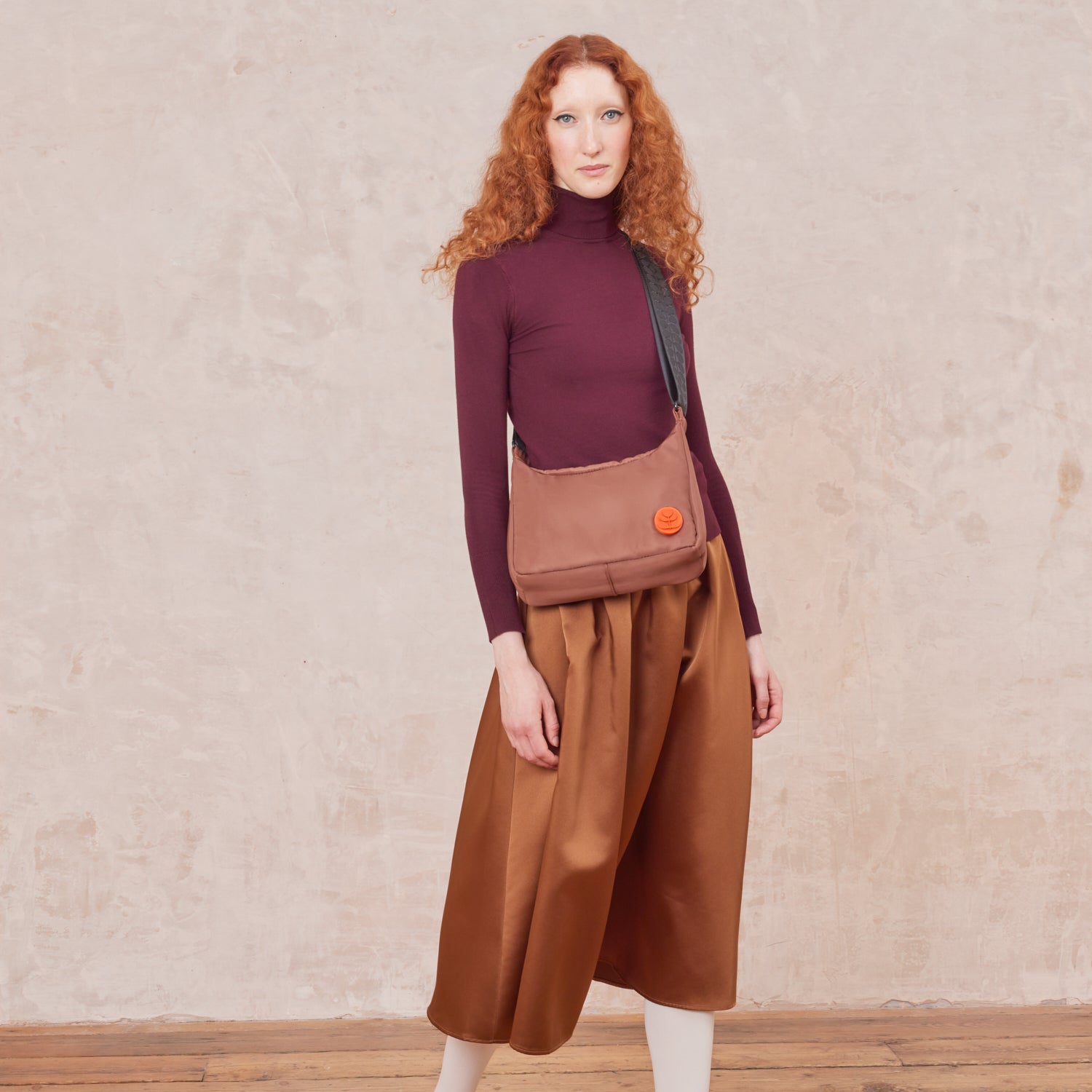 A women wears the Ratio Small bag in brown, with an adjustable crossbody strap. The handbag features an orange Orla Kiely logo.