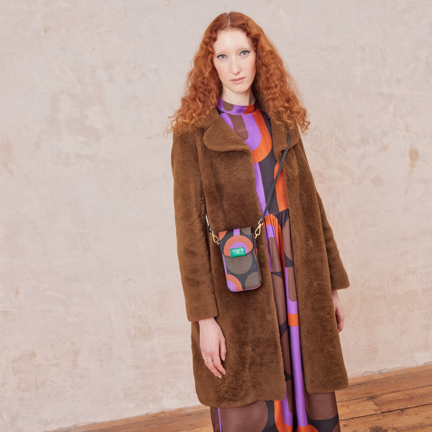 A woman wears a purple retro printed phone pouch across her body with a matching dress and brown winter coat.