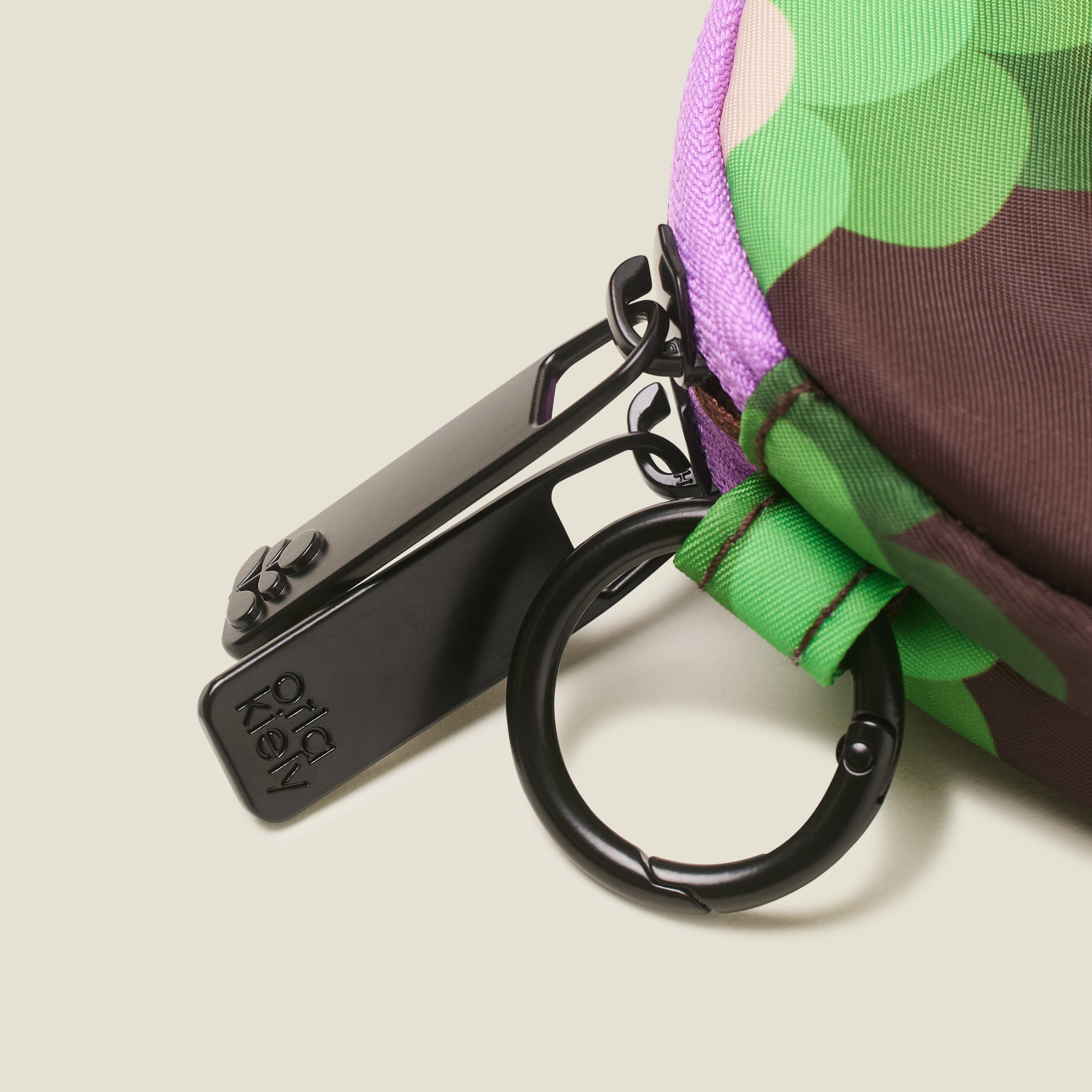 Close up of round change purse, in a green, brown and cream floral print with a ring clip and branded zip pulls on a beige background.