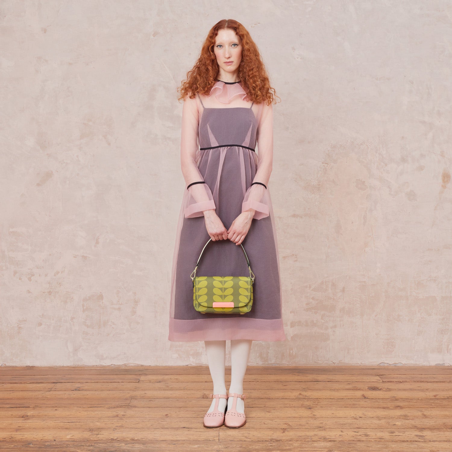 A woman in a pink dress holds a handbag with a green leaf pattern in front of her by the shoulder strap.