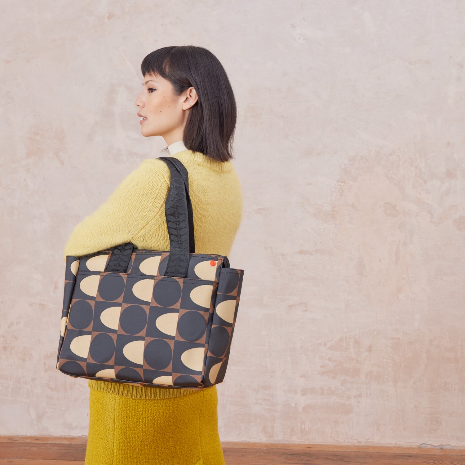 Woman carrying over her shoulder a brown, black and cream patterned tote bag with branded grab handles