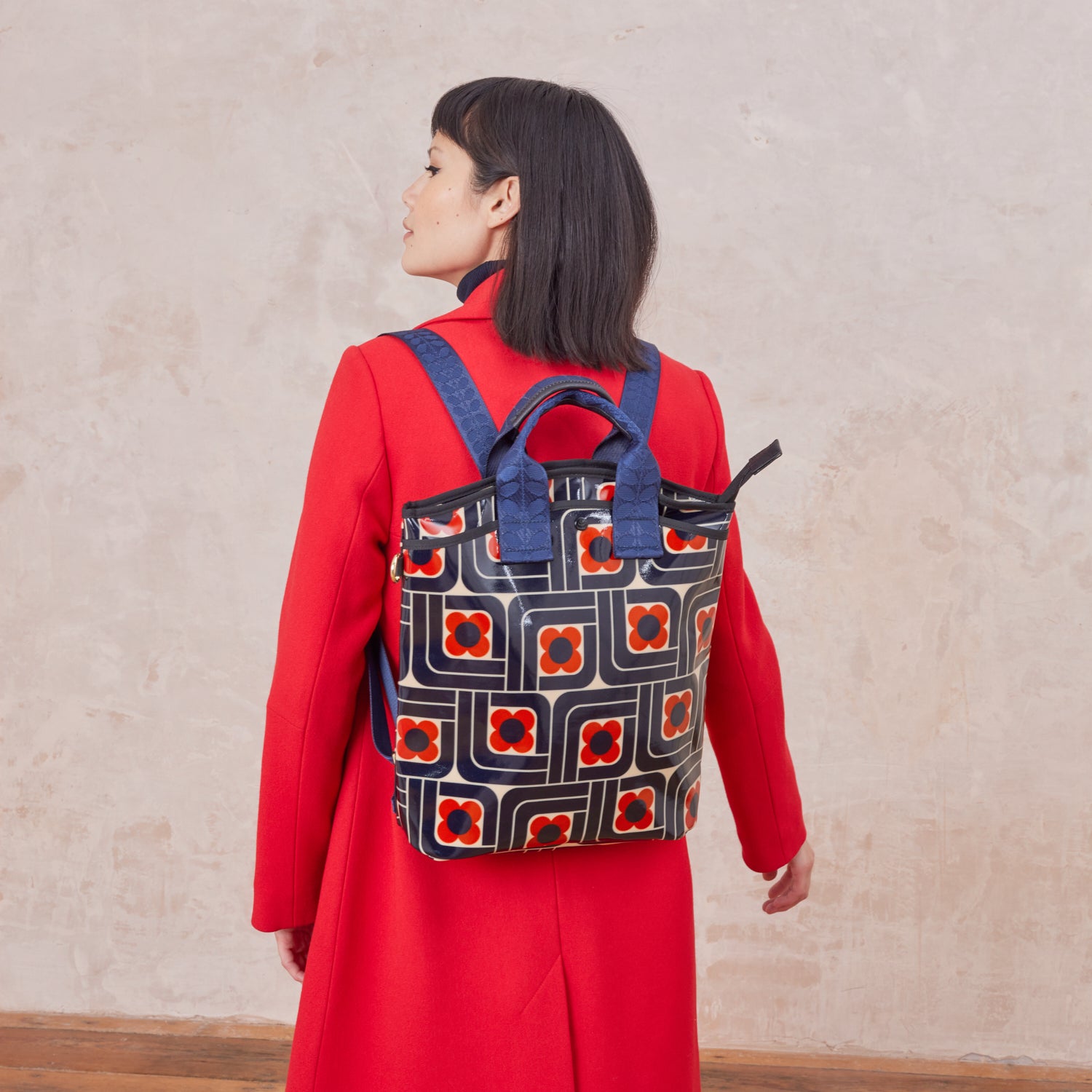 A woman wears a backpack in a navy blue and red geometric retro floral pattern in a red coat.