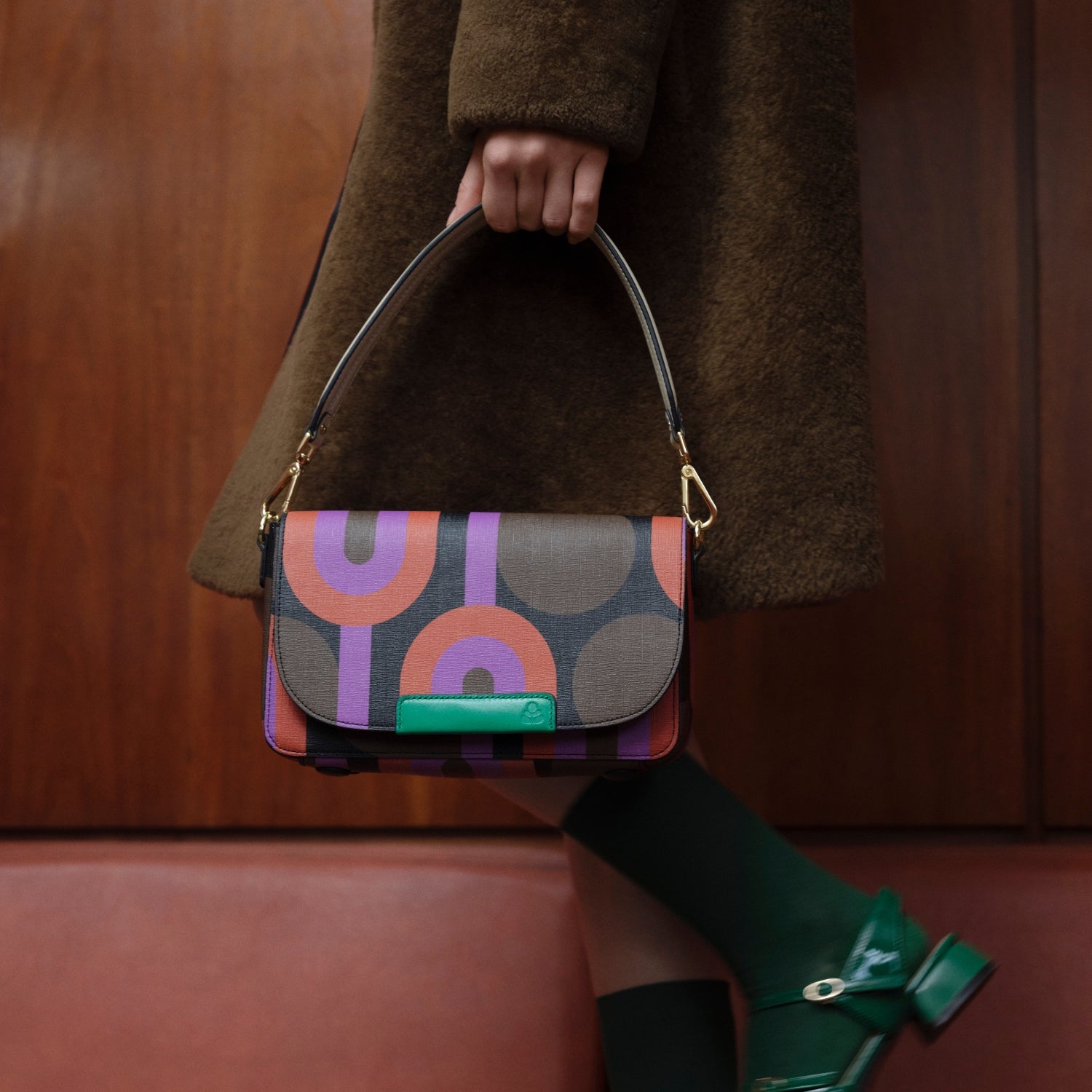 A woman holding a coloruful handbag with a patterned design, wearing a brown coat and green socks and shoes.