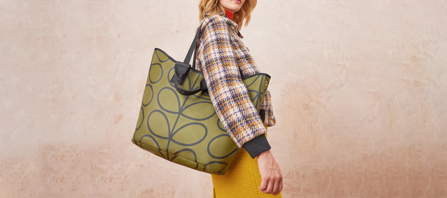 A women in a checked jacket wears a green shoulder bag by Orla Kiely in a grey stem print.