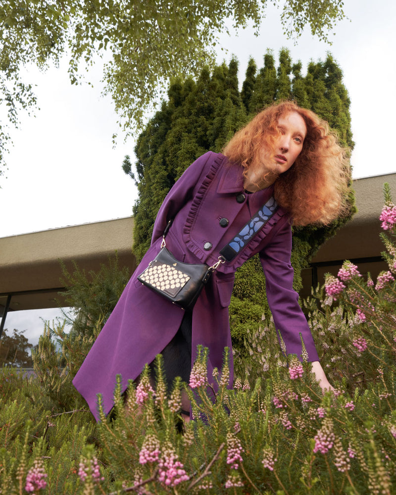 Woman standing amidst flowers wearing a floral leather Orla Kiely handbag