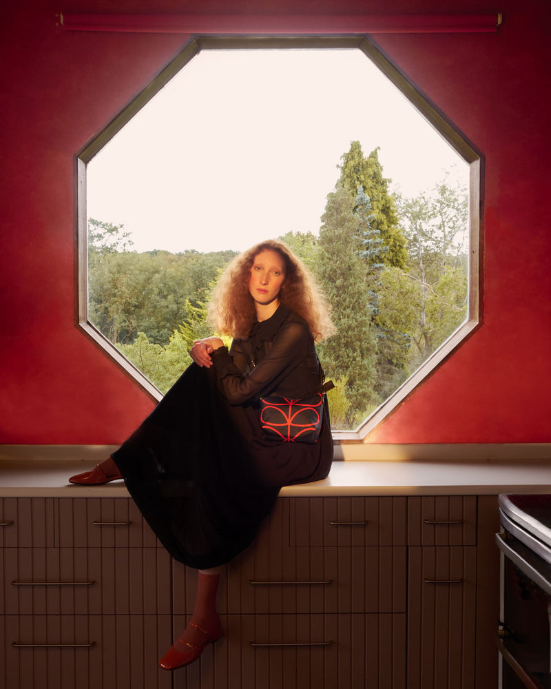 Model sitting by a hexagonal window wearing an Orla Kiely Carrymin Crossbody bag