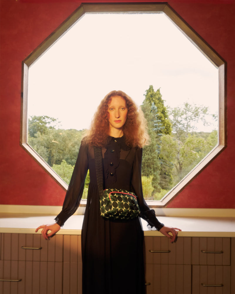 Model sitting by a hexagonal window wearing an Orla Kiely Angle Camera Bag