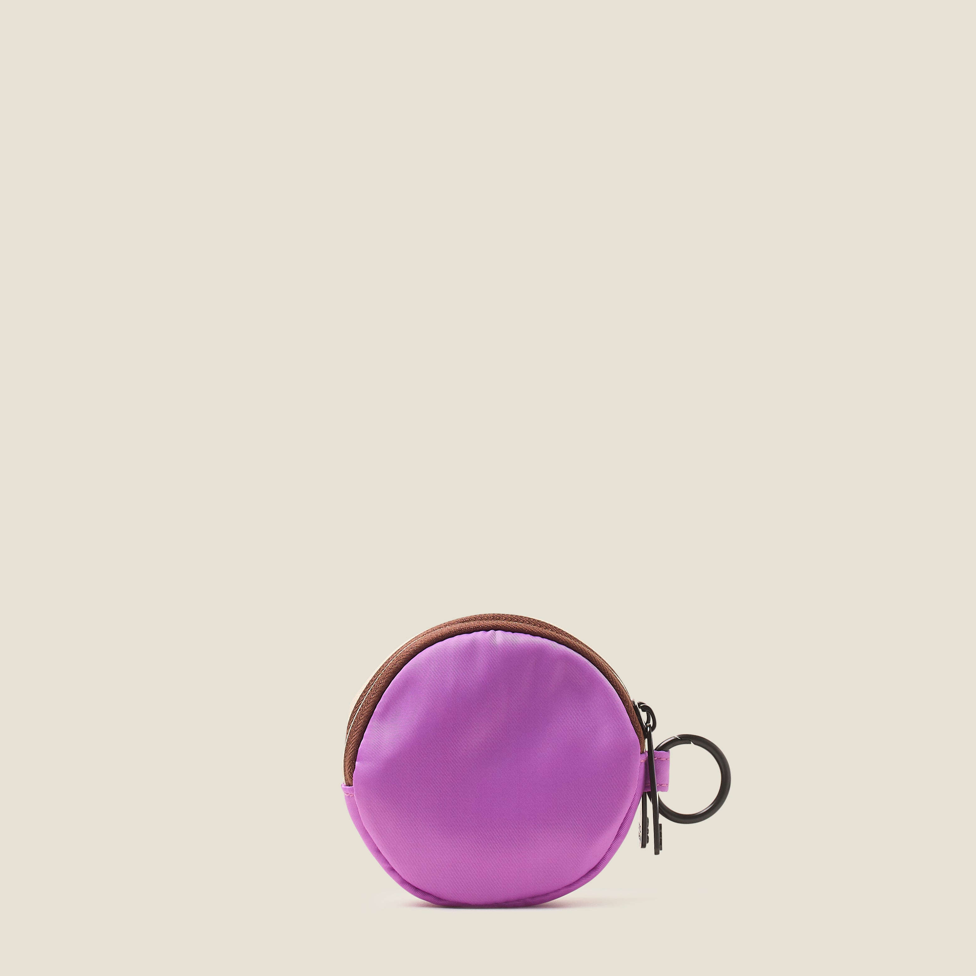 The back view of a lilac purple round coin purse with a rubber yellow Orla Kiely stem logo and ring clip on a beige background