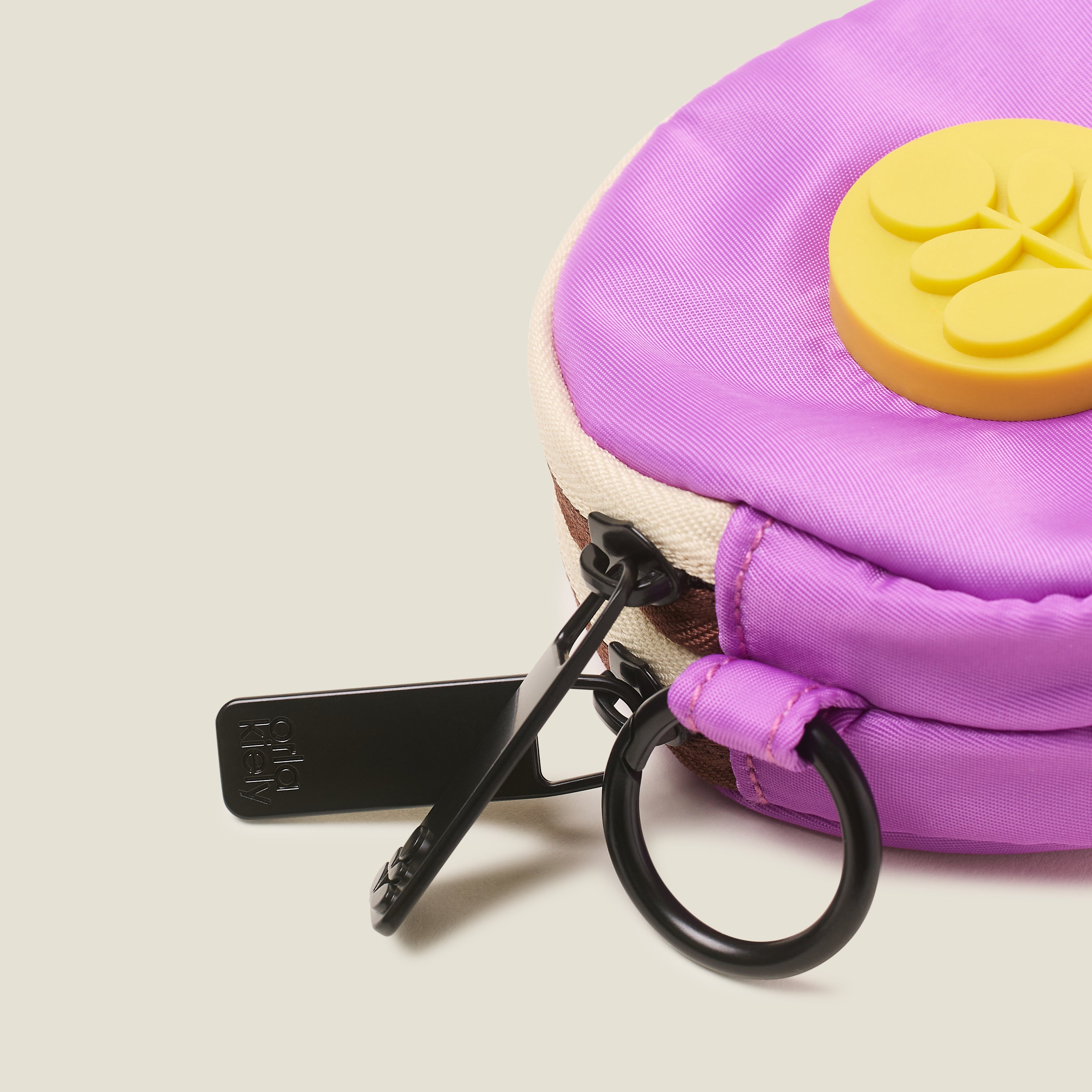 Close of view of a lilac purple round change purse with a rubber yellow Orla Kiely stem logo, ring clip and branded zip pulls on a beige background