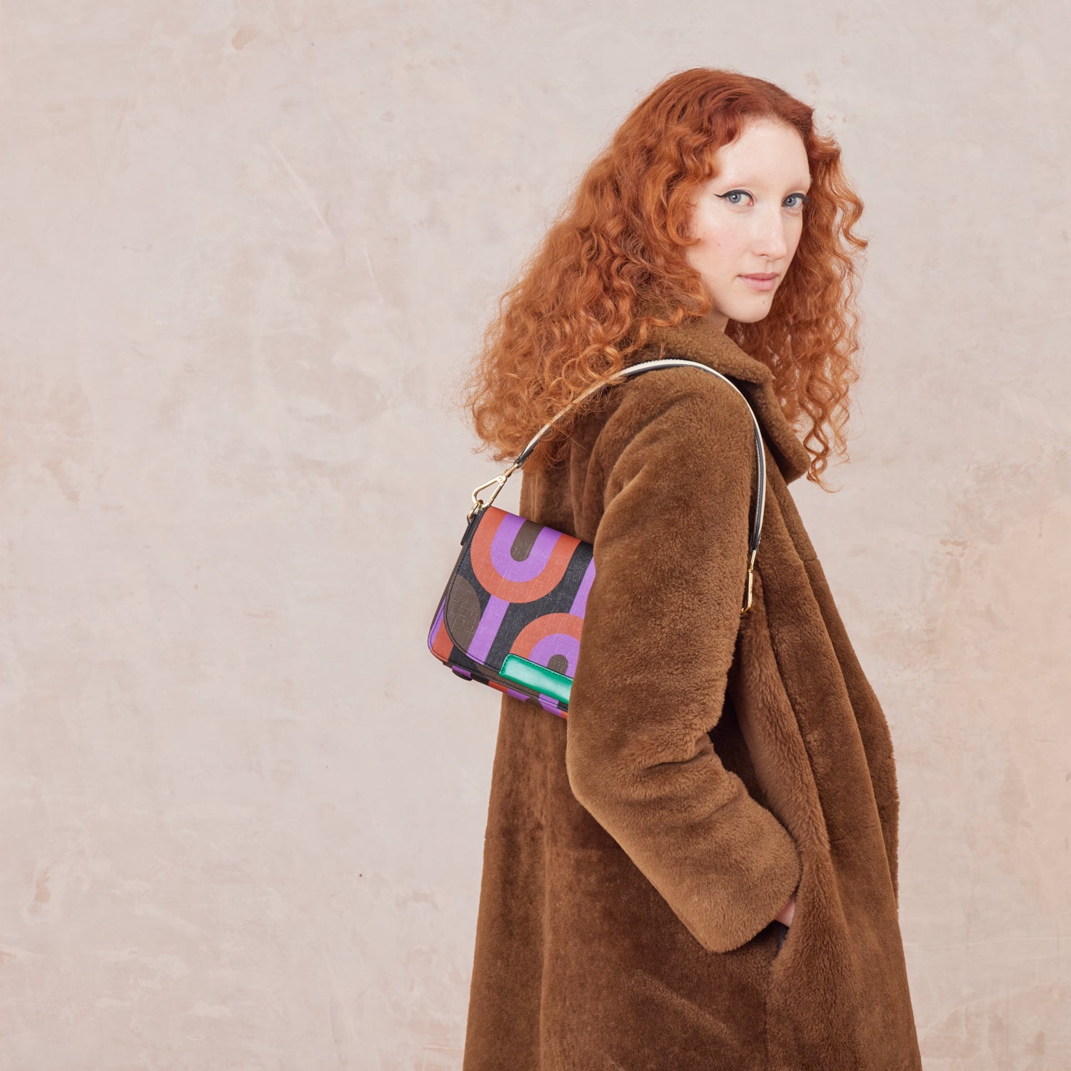 Woman in a brown coat wears a colourful  retro print handbag over her shoulder.