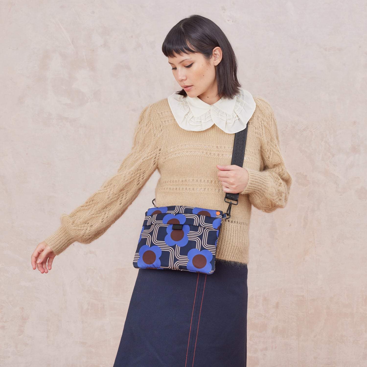 Woman wearing a beige sweater with a white collar and holding a blue patterned crossbody bag against a beige background