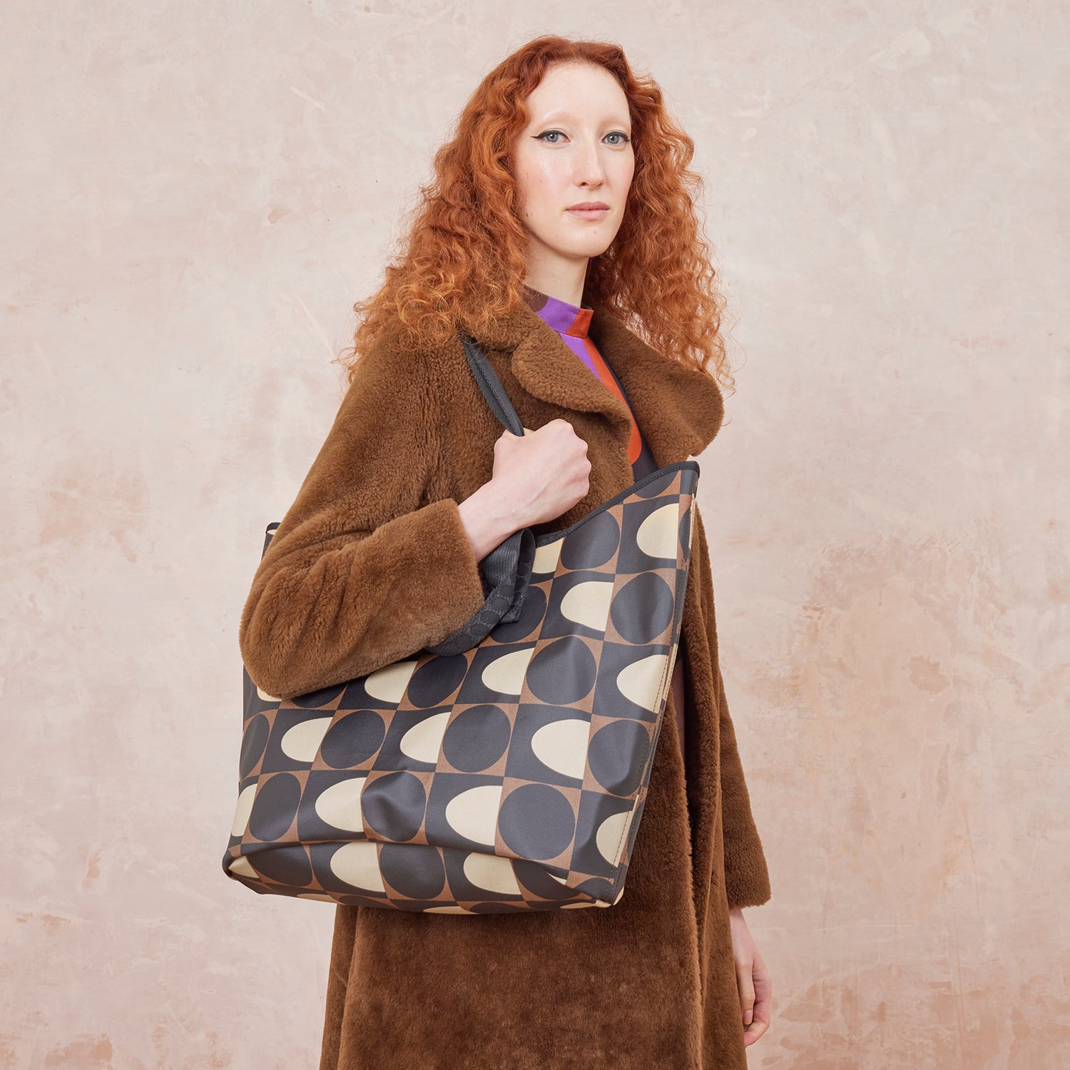 A woman wears a patterned tote bag over her sholuder with black, brown and beige spot square printed design.