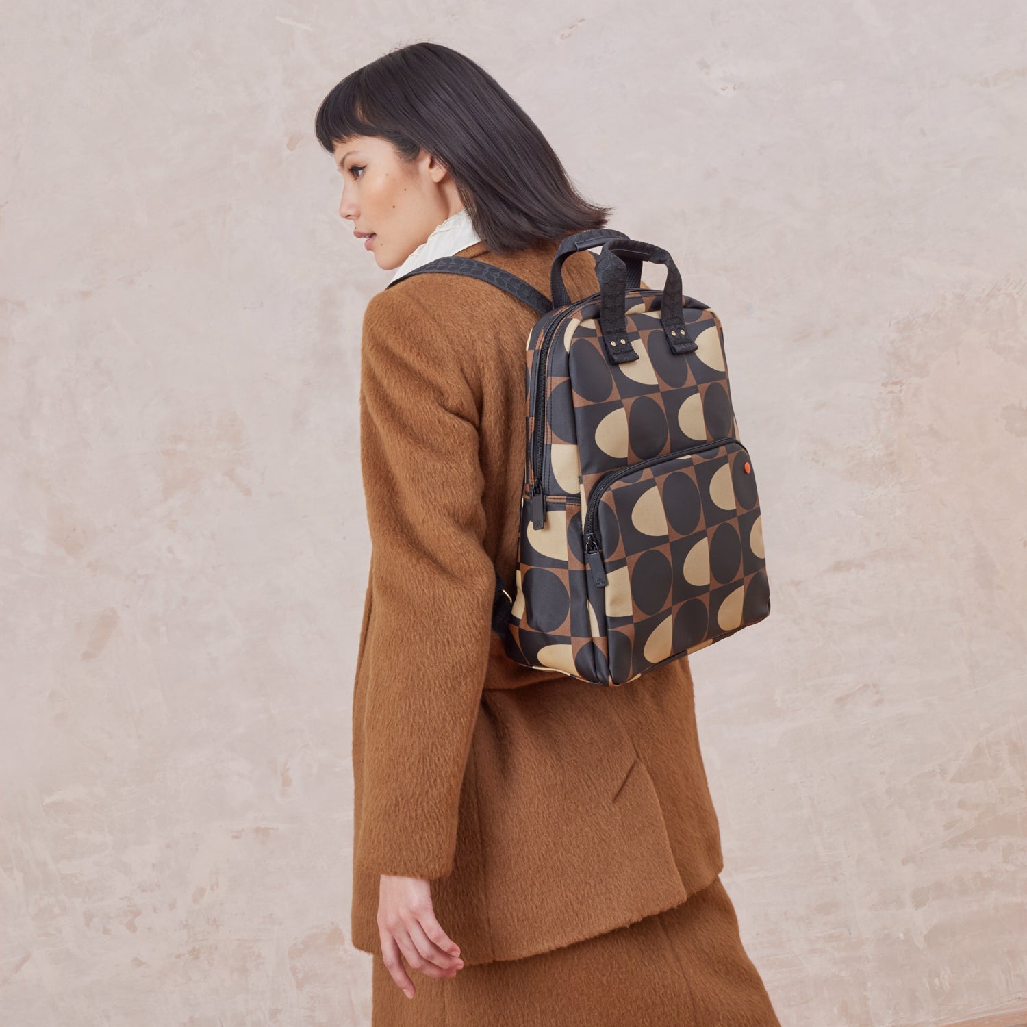 A women's wears the Lotta Backpack in a brown and cream dot and squares print featuring a zipped front pocket, branded grab handles and adjustable shoulder straps.