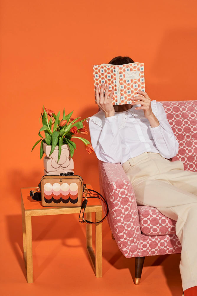 Person reading Little Women on an Orla Kiely pink sofa next to a leather bag 